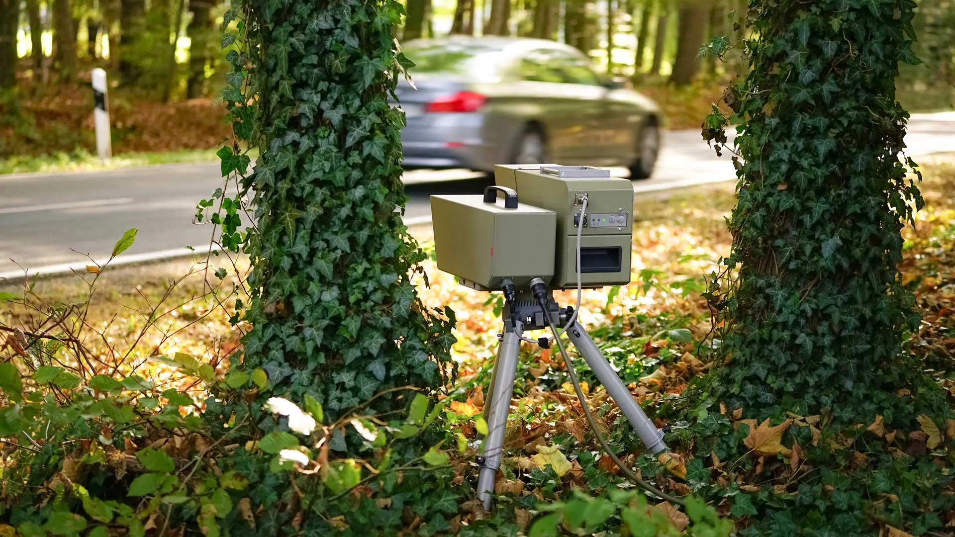 Das Bild zeigt einen mobilen Blitzer zwischen zwei Bäumen und ein vorbeifahrendes Auto.